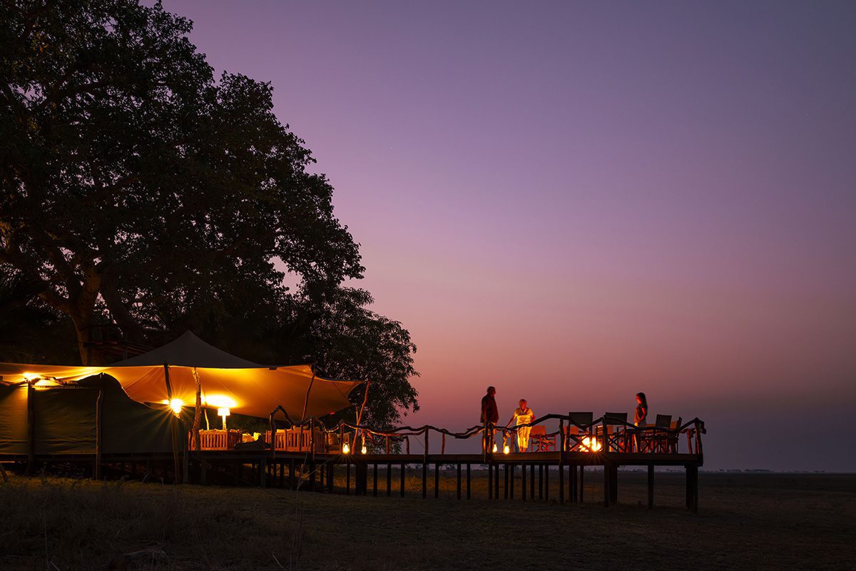 A safari camp at twilight, with a large illuminate tent and an elevated wooden deck, where people are gathered around a fire pit, under a vibrant purple sky.