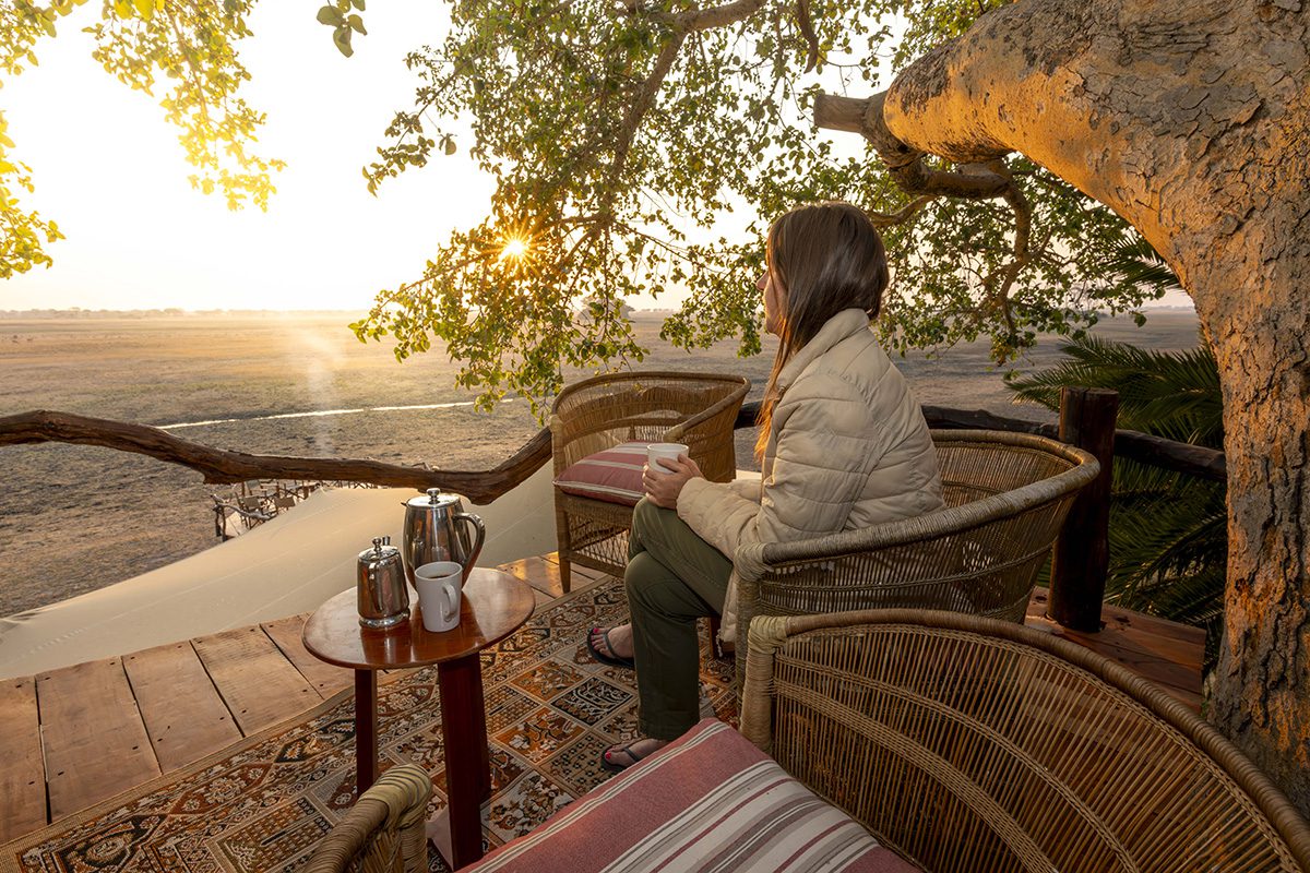 A side-on shot of a woman sat on a wooden deck, under a large tree, holding a mug, looking out at a vast landscape.
