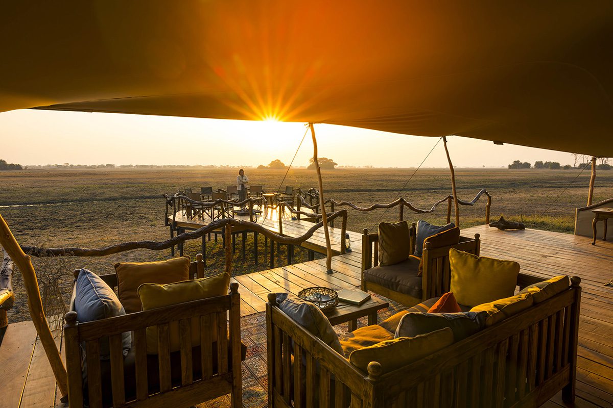 A golden sunset lights up a safari camp's wooden deck, featuring comfortable outdoor seating, under a canvas canopy, with a flat landscape stretching into the horizon.
