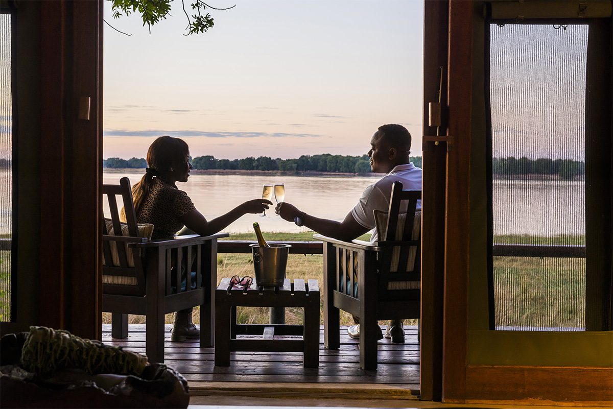 A couple sat on a wooden deck, raising a toast, whilst looking out at a wide river in front.