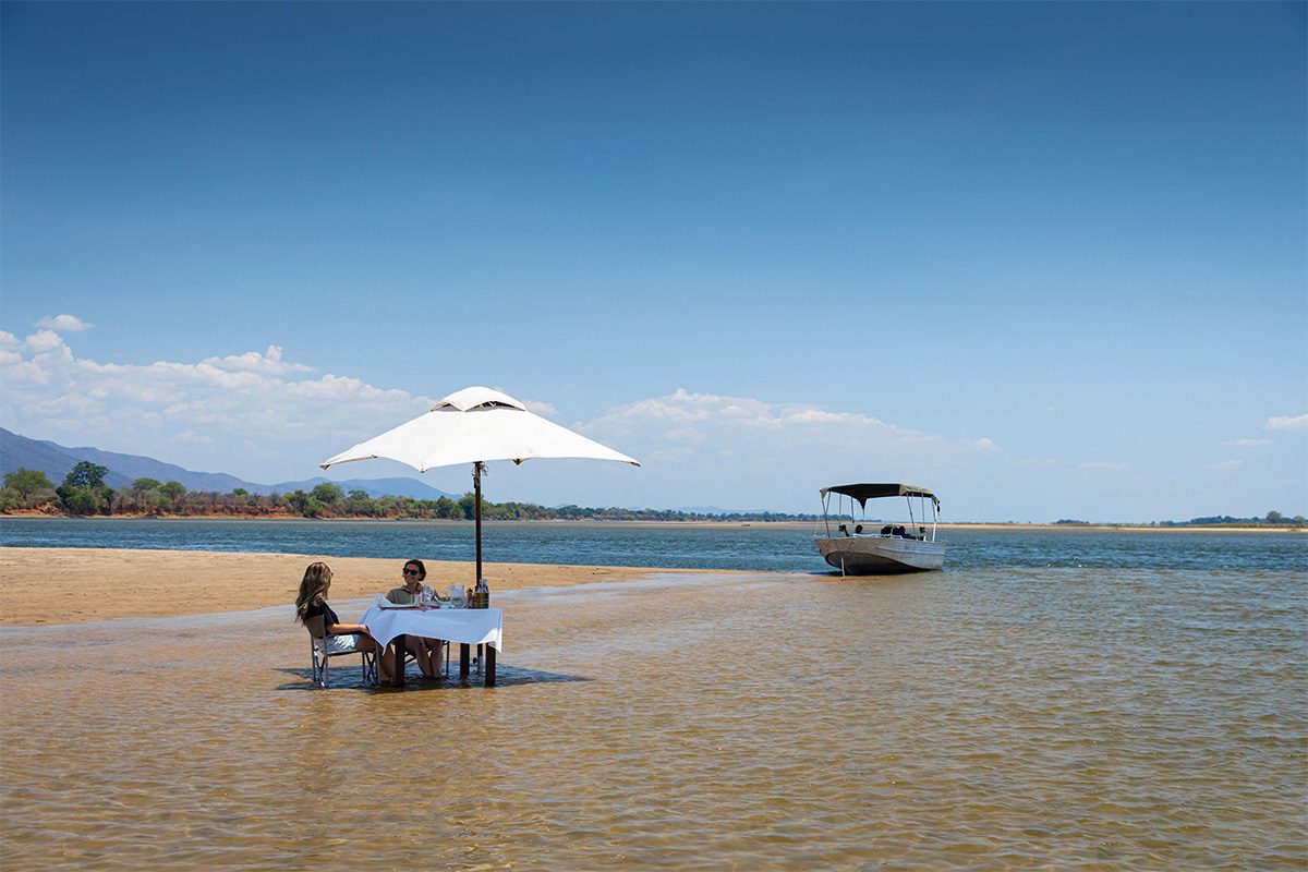 Two people situated at a white table with an umbrella set up, sat in shallow water, on what appears to be a sandbank.