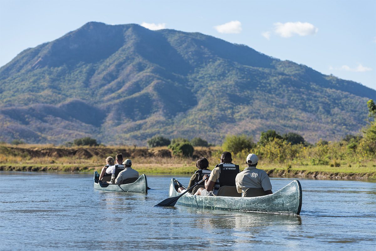 Two full canoes paddling down a calm river, with a large mountain in the background, under a blue sky.