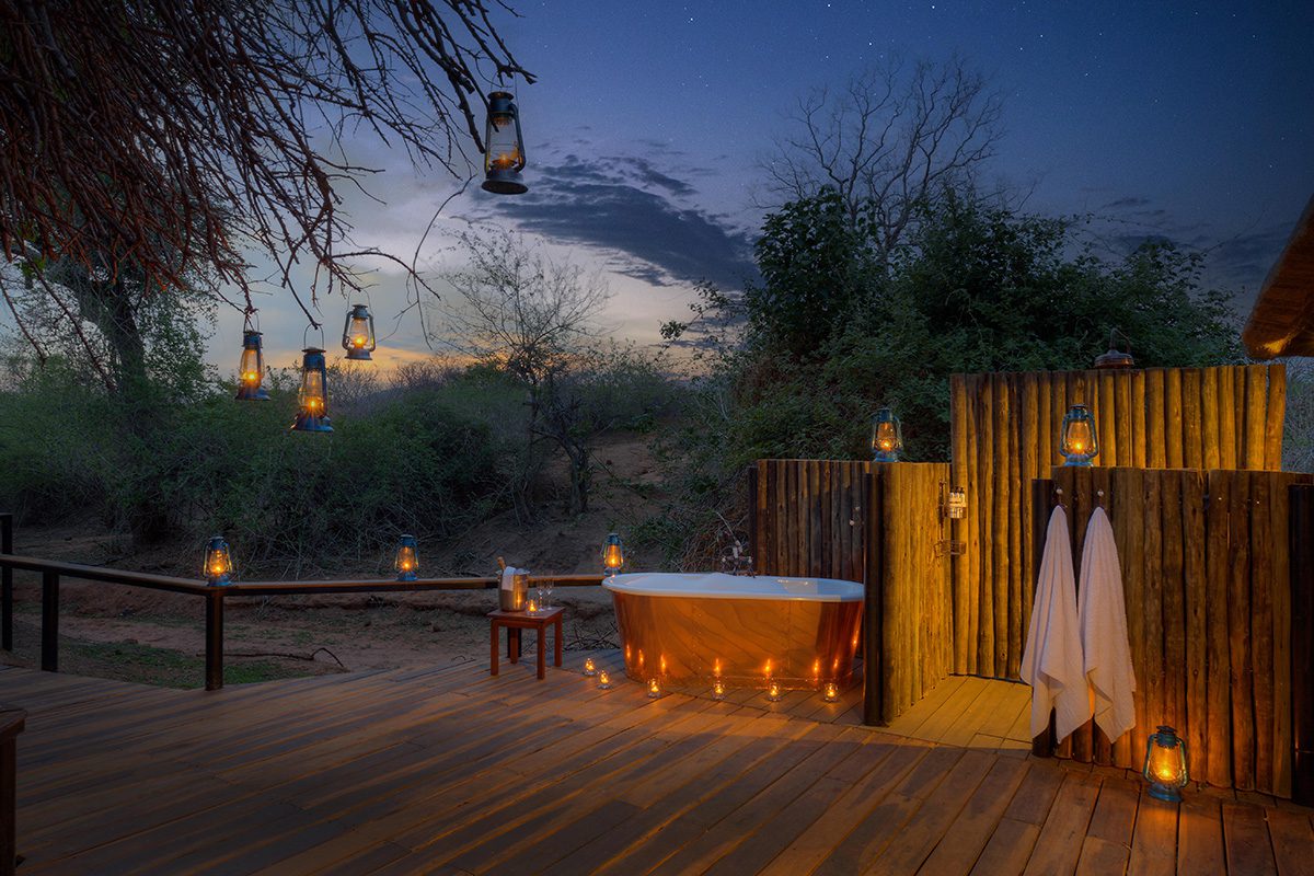 An outdoor bathroom setup at dusk, equipped with a freestanding bath tub and shower on a wooden deck. Surrounded by trees, the setup is illuminated by numerous lanterns and candles.