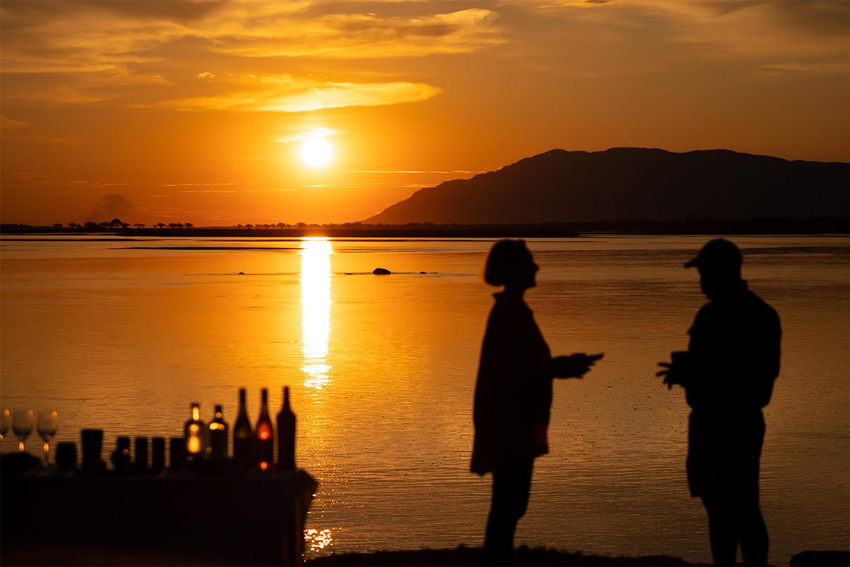 Two silhouettes of people stood by a body of water at sunset, with a table of drinks set up in the foreground.