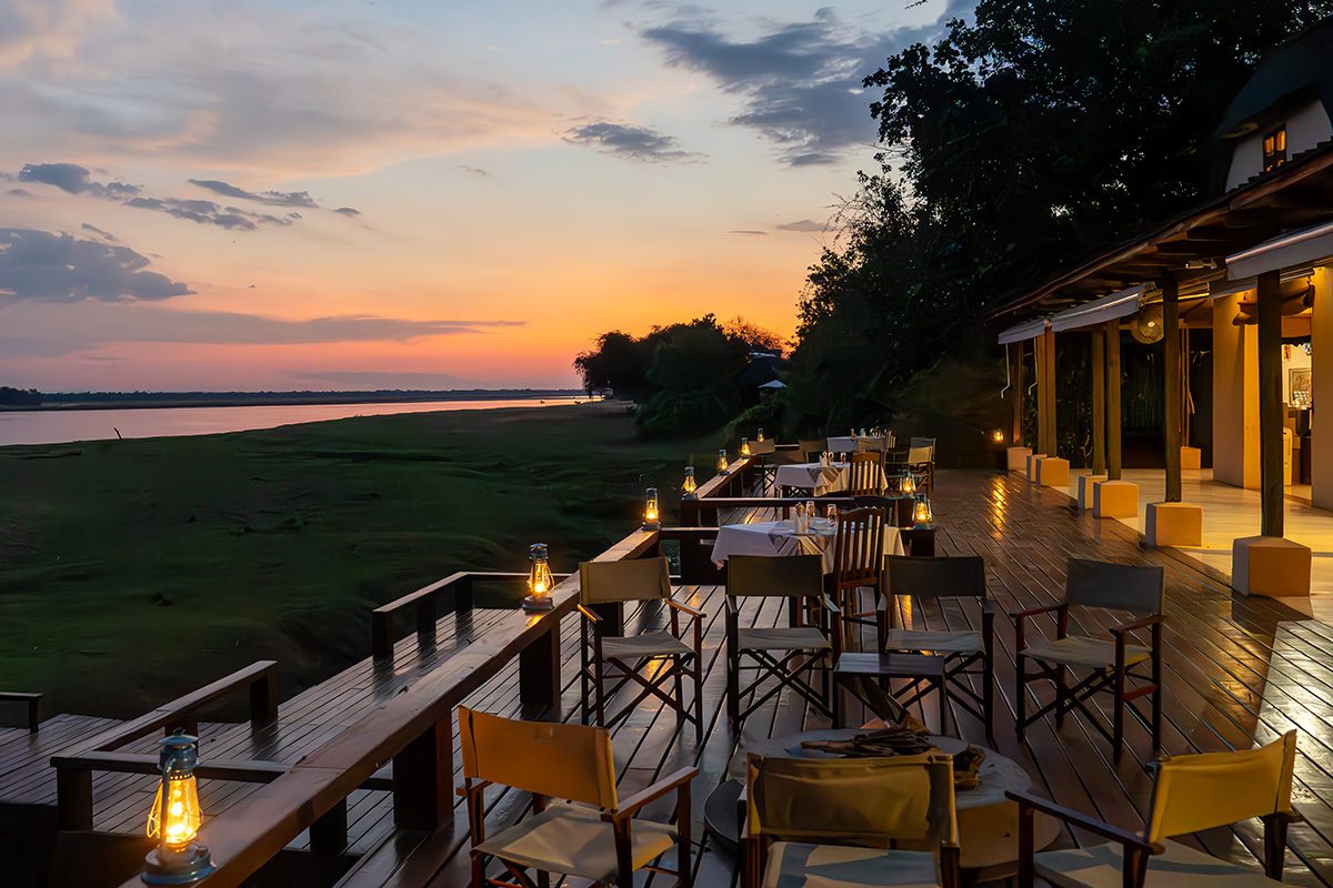 An outdoor dining deck at dusk, overlooking a wide river and grass bank. Chairs and tables are illuminated by lanterns, and the warm glow of the sunset sky.