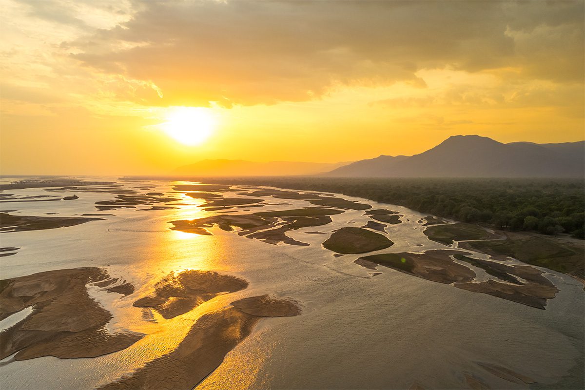 An aerial view of a wide river filled with numerous sandbanks and small islands, reflecting the bright light of a sunset, with dark mountains in the distance.