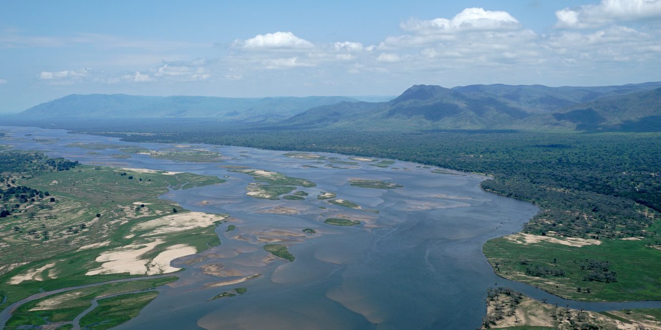 An aerial view of a wide river with many sandy islands and a large expanse of water. The river is surrounded by lush green landscapes and a range of mountains in the distance under a blue sky with scattered clouds.