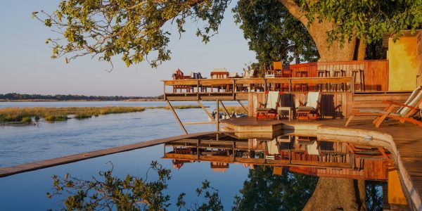 An infinity pool and a wooden deck with lounge chairs and dining tables overlook a river at dusk. The water in the pool reflects the sky and the trees.