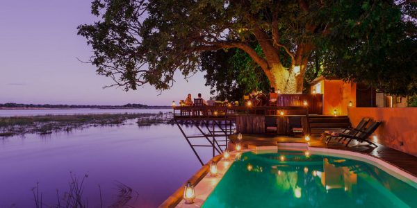 An infinity pool and a wooden deck with lounge chairs and dining tables overlook a river at dusk. The water in the pool reflects the sky and the trees.