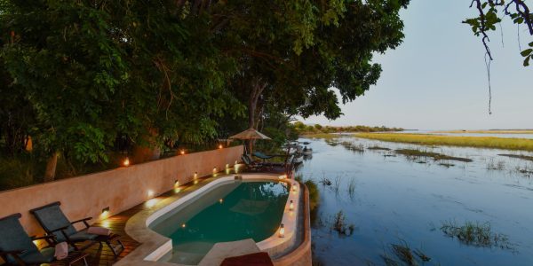 An outdoor infinity pool with lights surrounding at dusk, overlooking a river and a marshy landscape. The pool deck has lounge chairs, and is surrounded by trees.
