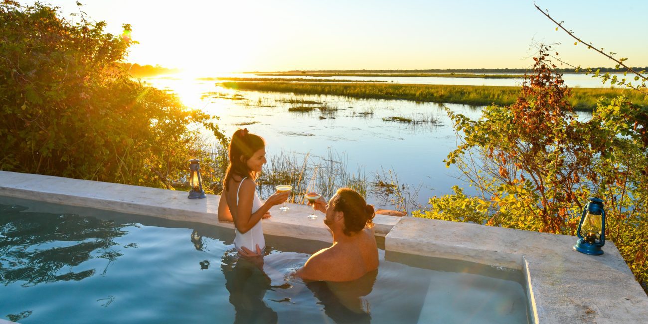 A couple is in their plunge pool, holding glasses and toasting, as the sun sets over a marshy landscape.