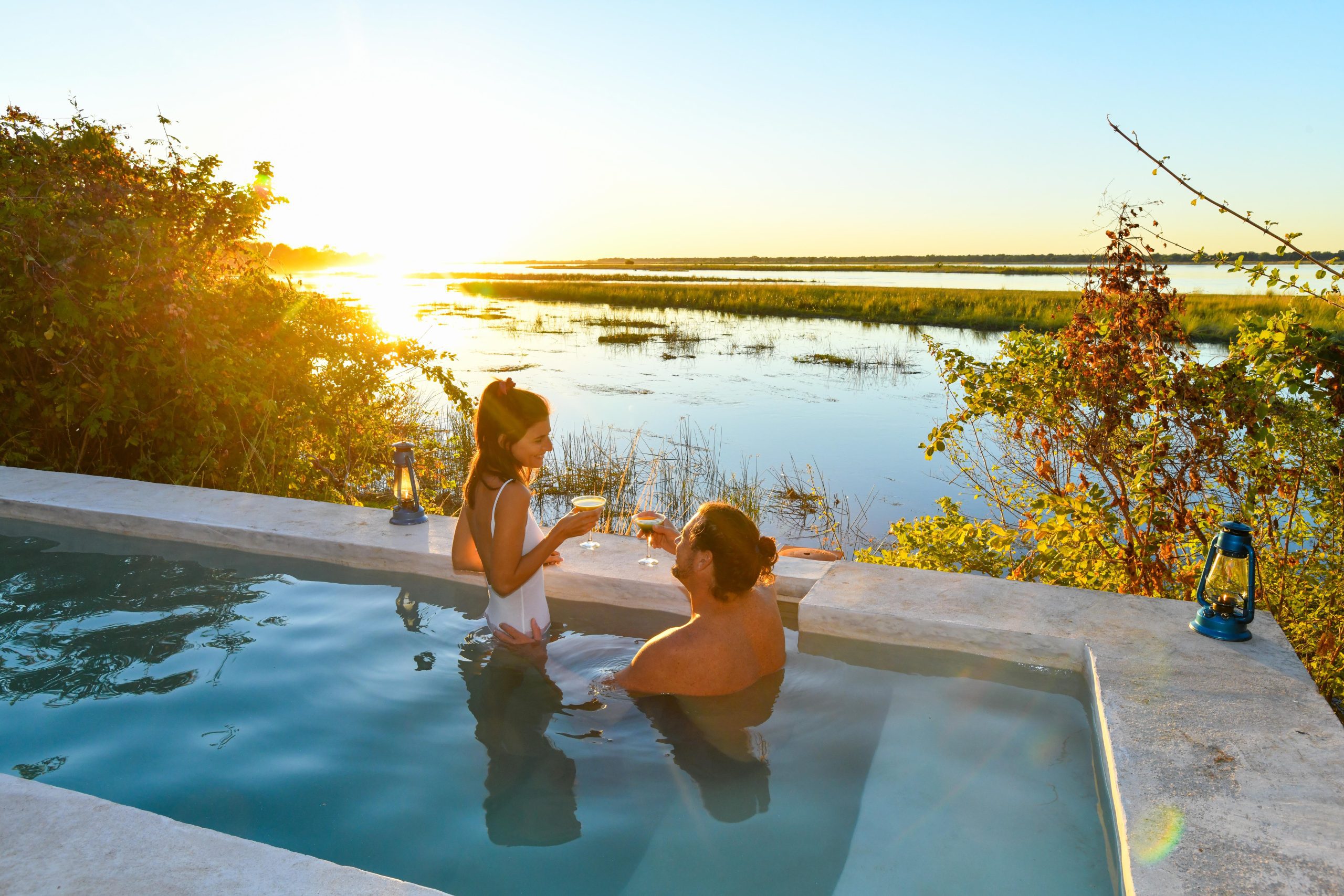 A couple is in their plunge pool, holding glasses and toasting, as the sun sets over a marshy landscape.