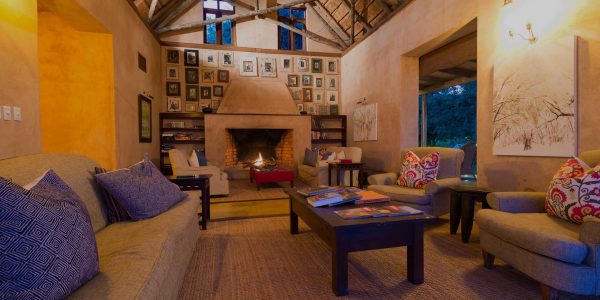 The interior of a cozy guest area features a high, thatched roof, and a large fireplace. The room is decorated with comfortable sofas and chairs, and a gallery wall of framed photos hangs above the fireplace.